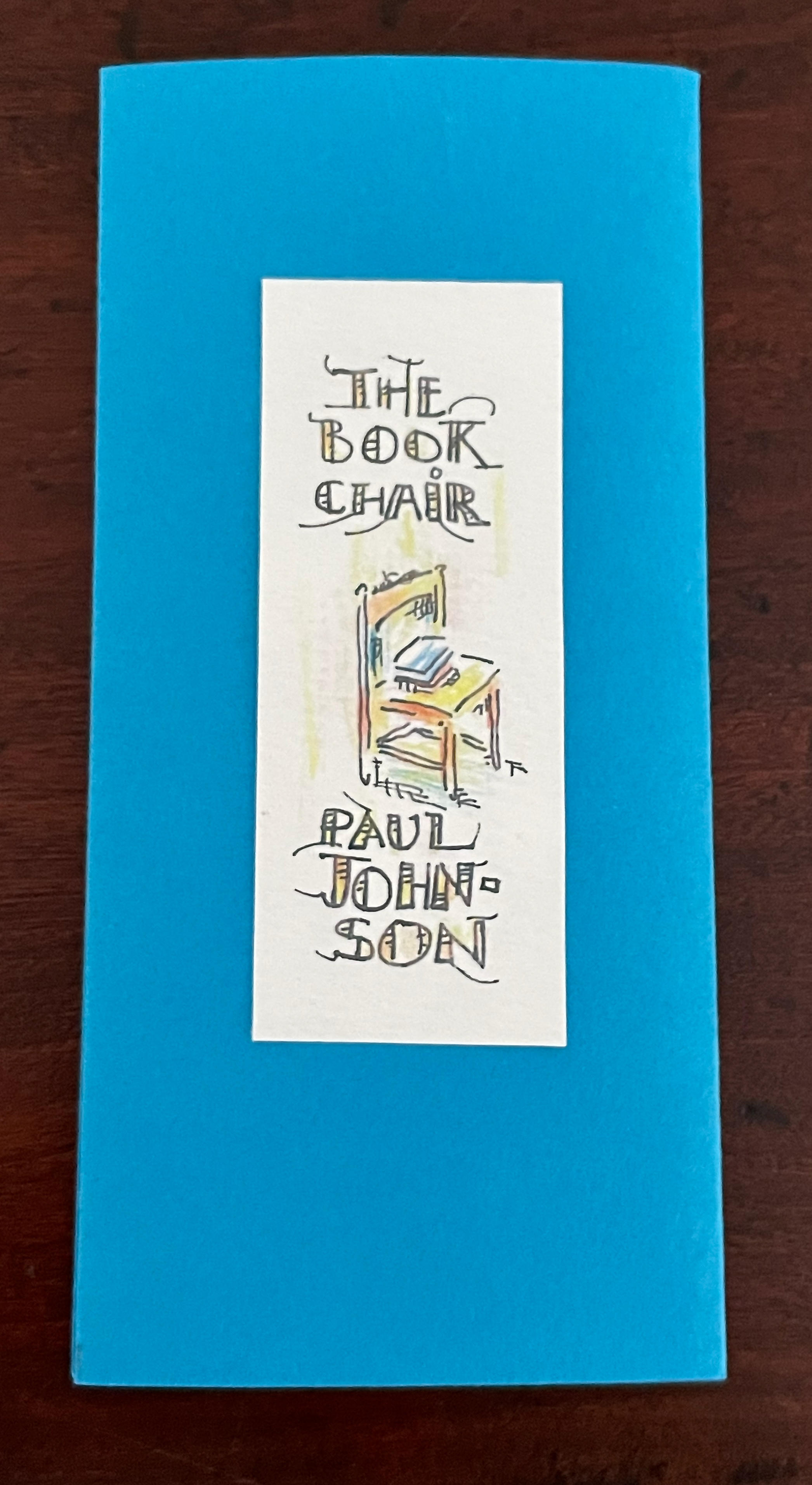 A blue booklet cover with an illustrated label featuring the title 'The Book Chair' and the author's name, 'Paul Johnson.' The label includes a sketch of a chair and colorful accents.