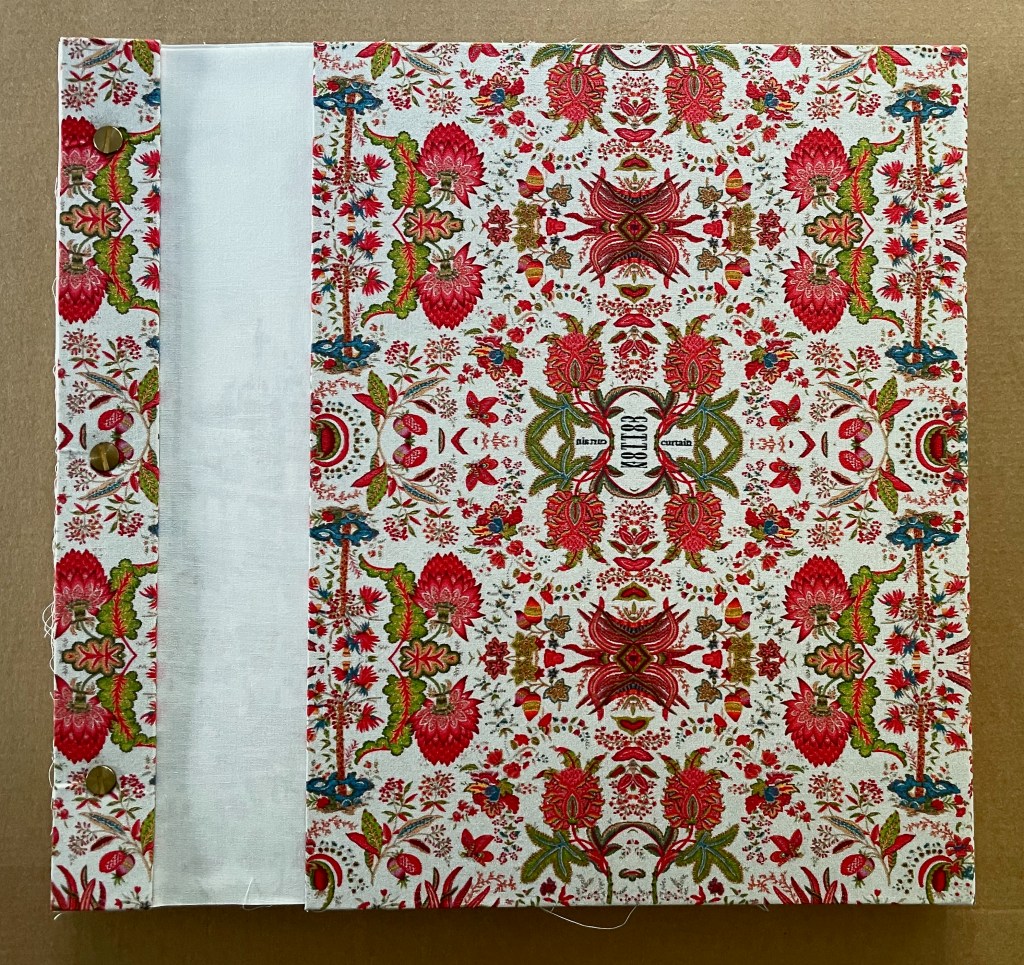 A decorative cover featuring a floral pattern in red, green, and blue on a white background, with a plain white spine and two brass buttons.