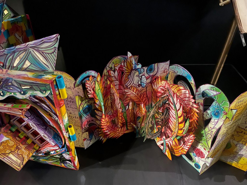 A colorful pop-up artwork featuring intricate designs, including flowers, leaves, and abstract shapes, set against a black background.