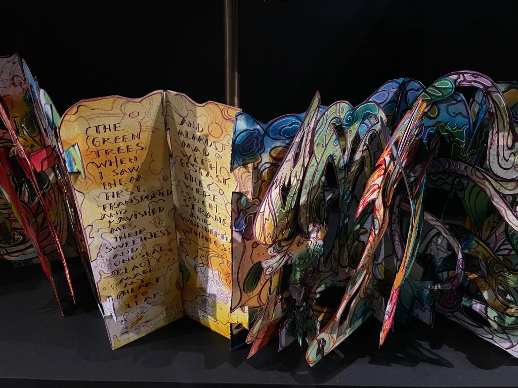 An artistic pop-up book featuring colorful, intricate designs and handwritten text, prominently displaying the phrase 'The green trees when I saw them...' within a layered, three-dimensional format.