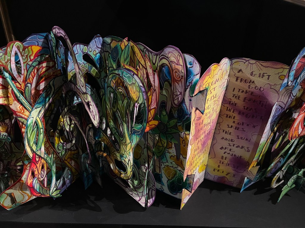 A colorful paper art sculpture featuring intricate patterns and designs, with portions of handwritten text visible among the vibrant decorations.