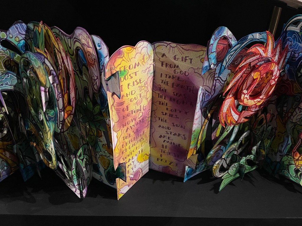 Colorful pop-up art piece featuring intricate designs and poetry, with phrases about rising from dust and a gift from God.