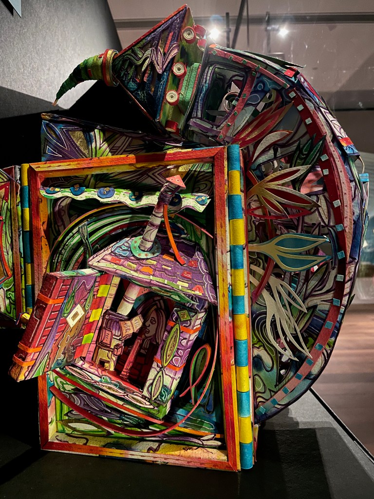 A colorful, intricate three-dimensional paper sculpture featuring various abstract shapes, designs, and vibrant patterns, resembling a whimsical house with plants and exaggerated architectural elements.