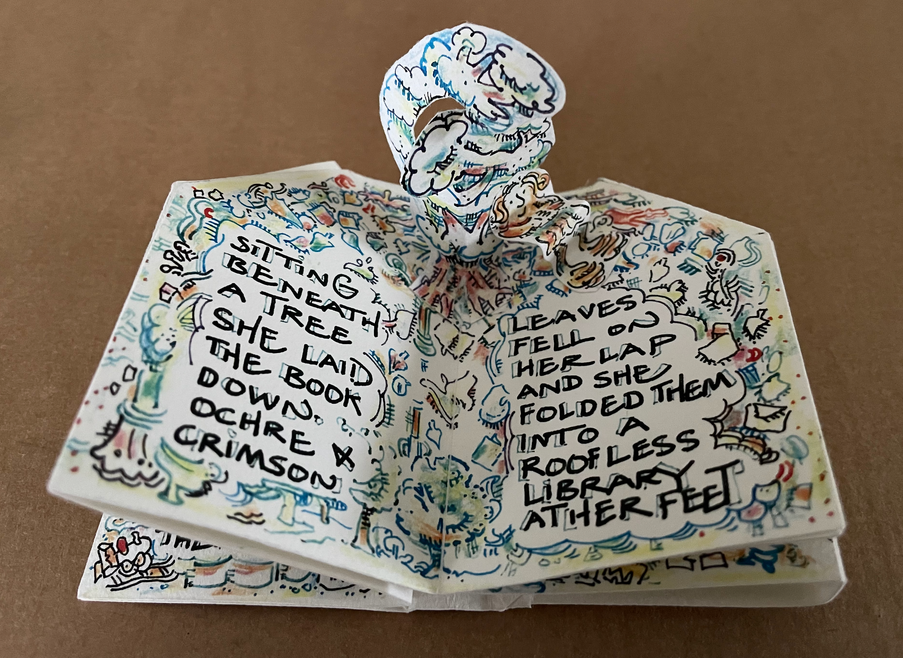 A pop-up art book open to two pages with colorful illustrations and handwritten text. The top page features a pop-up figure surrounded by whimsical drawings, while the text includes phrases about sitting beneath a tree and leaves falling into a lap.