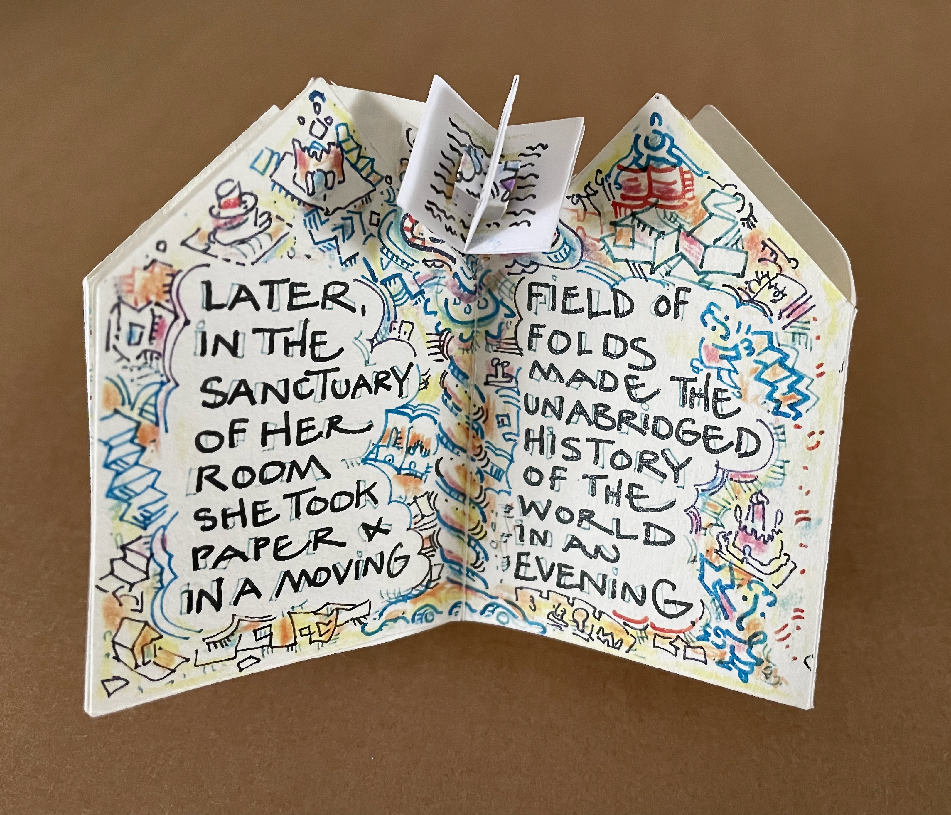 Open handmade book with colorful illustrations and handwritten text: 'Later, in the sanctuary of her room she took paper & in a moving field of folds made the unabridged history of the world in an evening.'