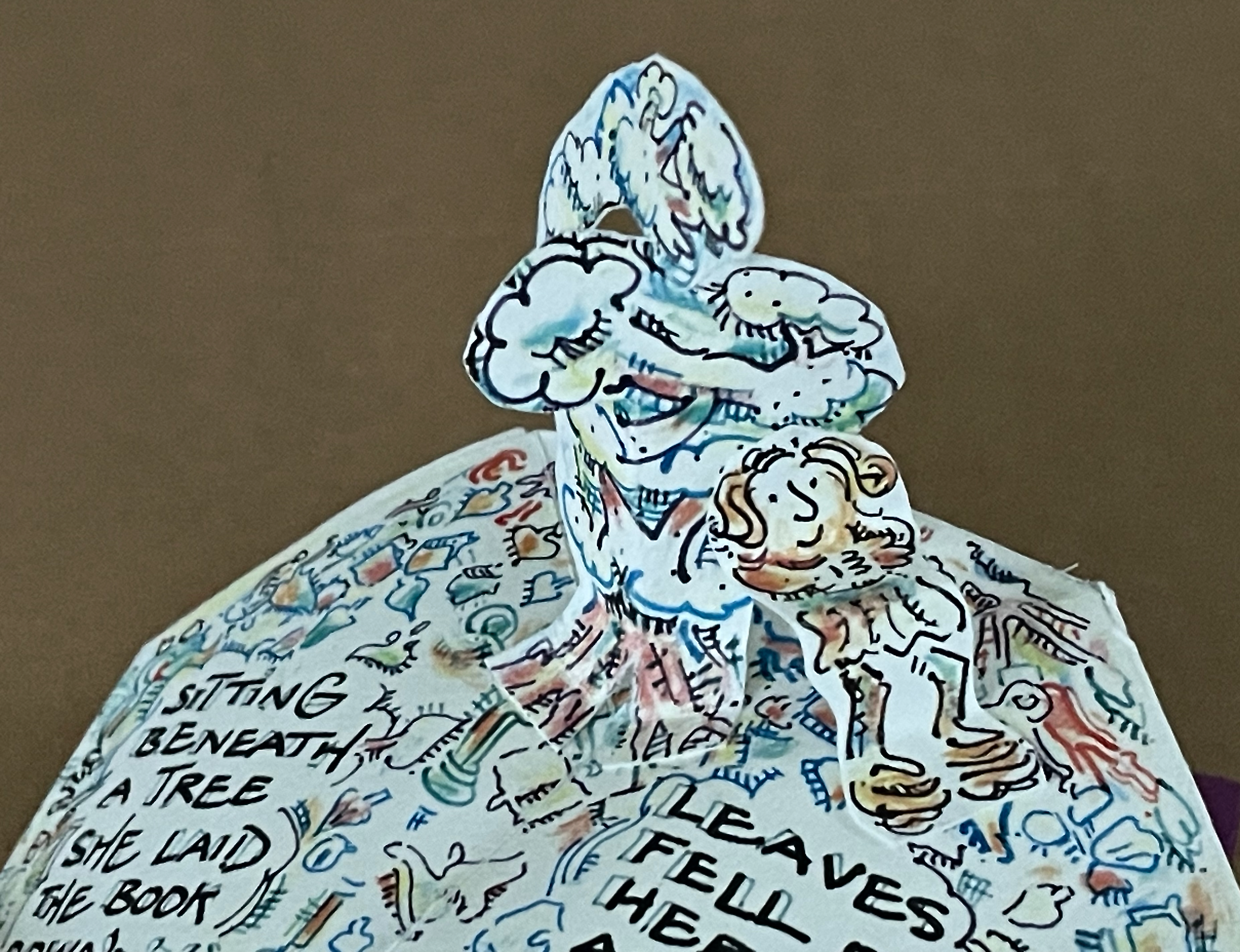 A pop-up book illustration depicting a colorful scene with a character sitting beneath a tree, surrounded by whimsical clouds and text that includes phrases about laying a book and falling leaves.