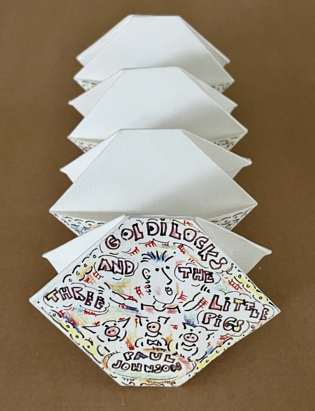 A set of folded paper pages with illustrations and text on the front cover that reads 'Goldilocks and the Three Little Pigs' by Paul Johnson.