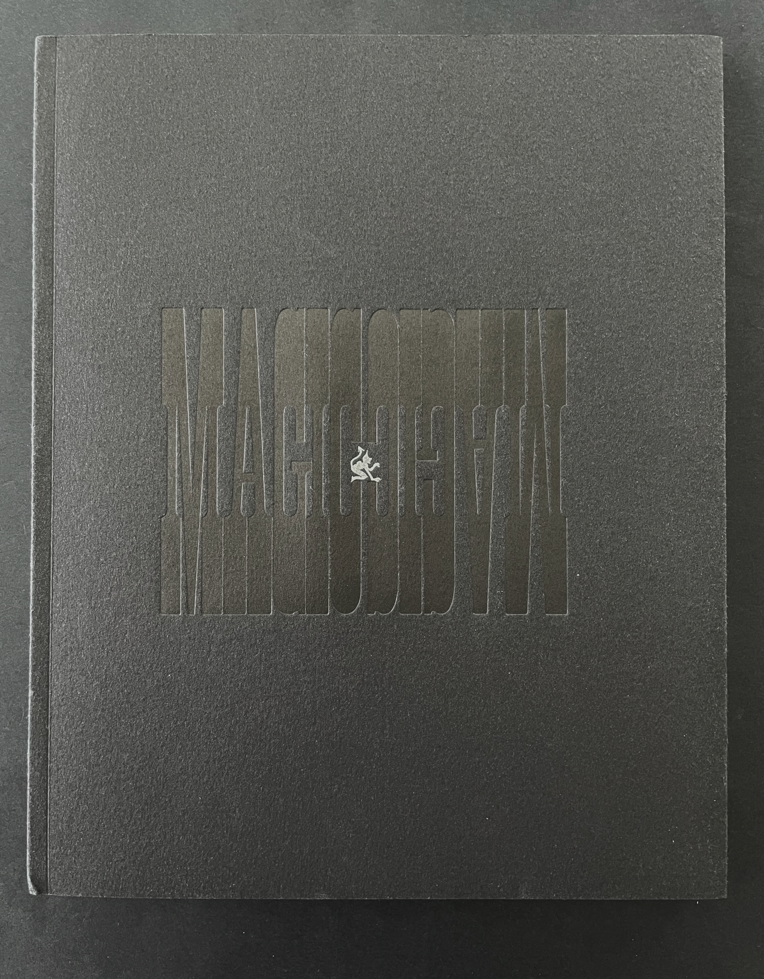 A black book cover with the title 'MAGMOODVIN' embossed in a lighter shade of black, featuring a small white logo or illustration centered at the bottom.