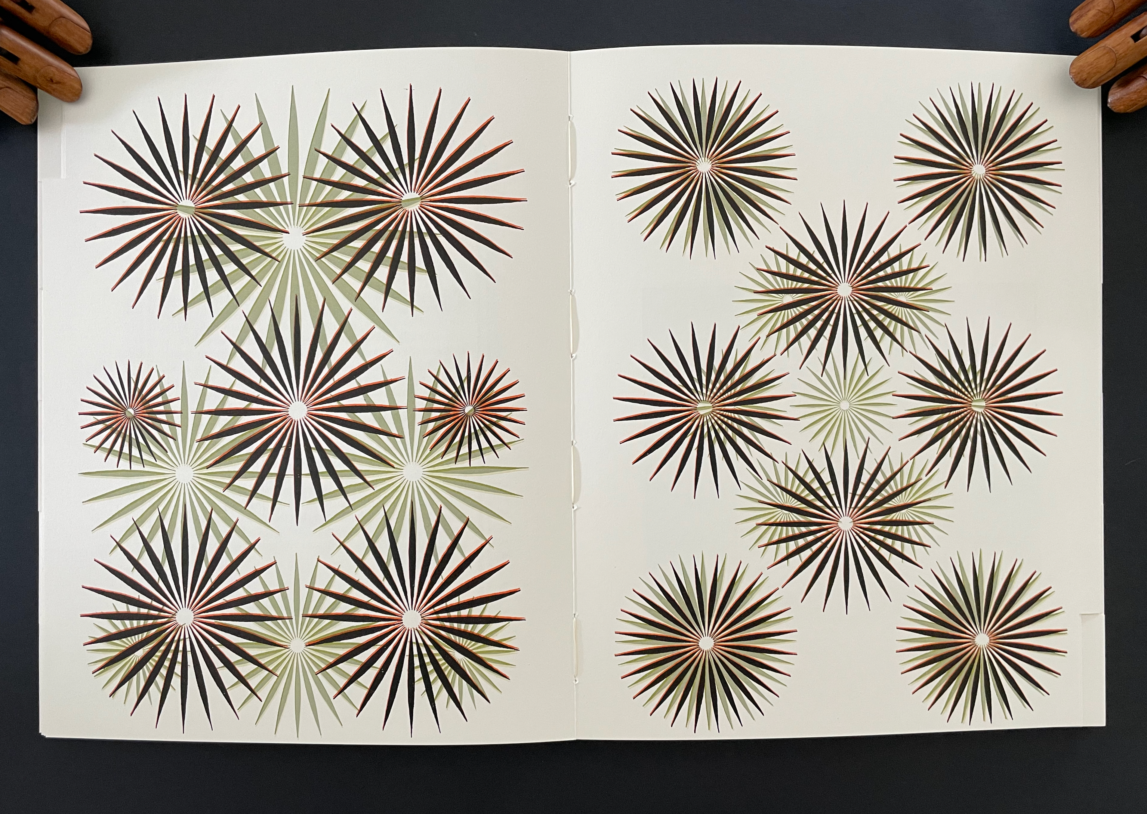 An open book displaying a page with intricate geometric patterns of starburst shapes in various colors, including green, black, and red on a cream background.