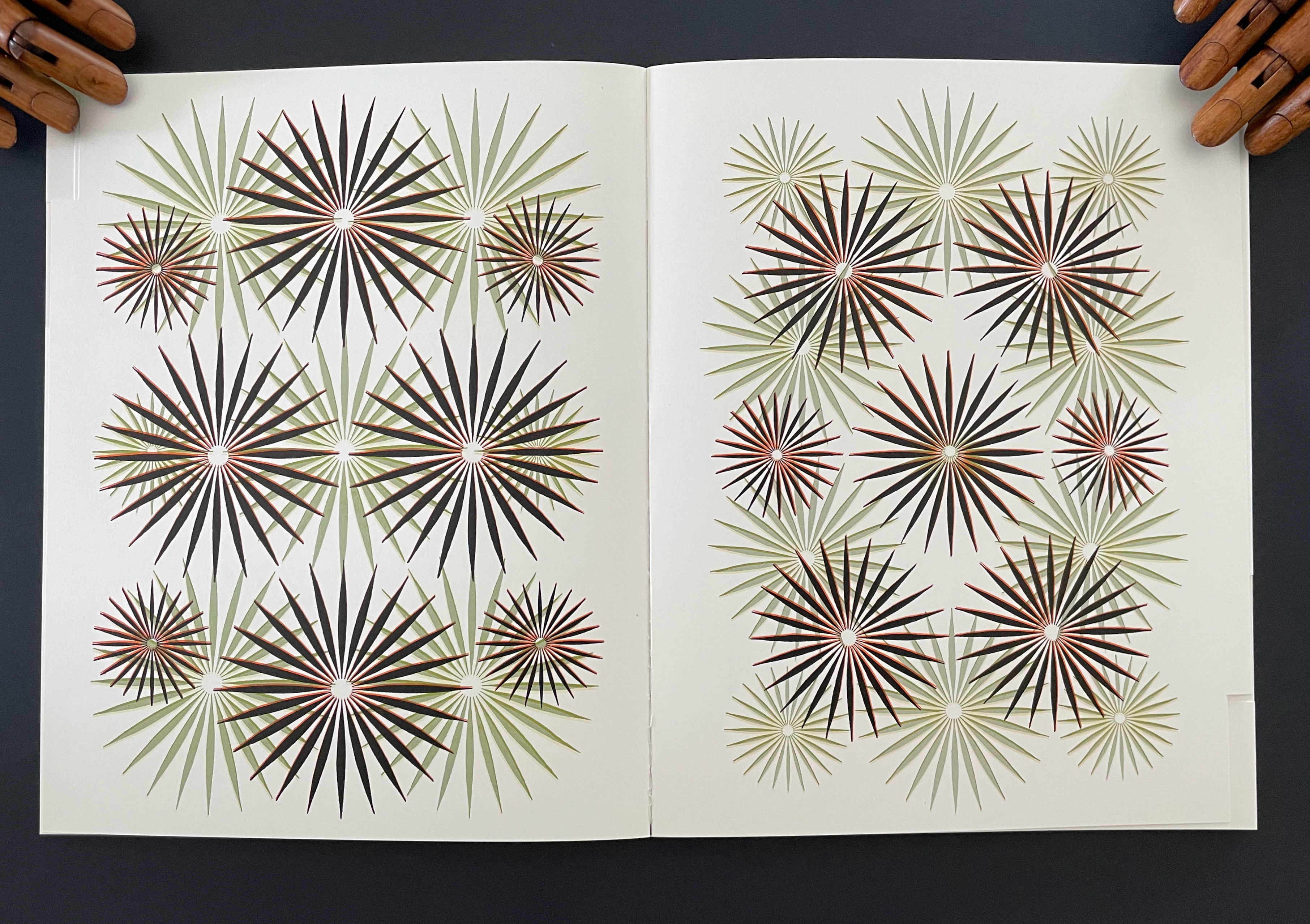 A symmetrical design featuring intricate cut-out patterns of star-like shapes in various colors including black, green, and red, displayed on an open book against a dark background.