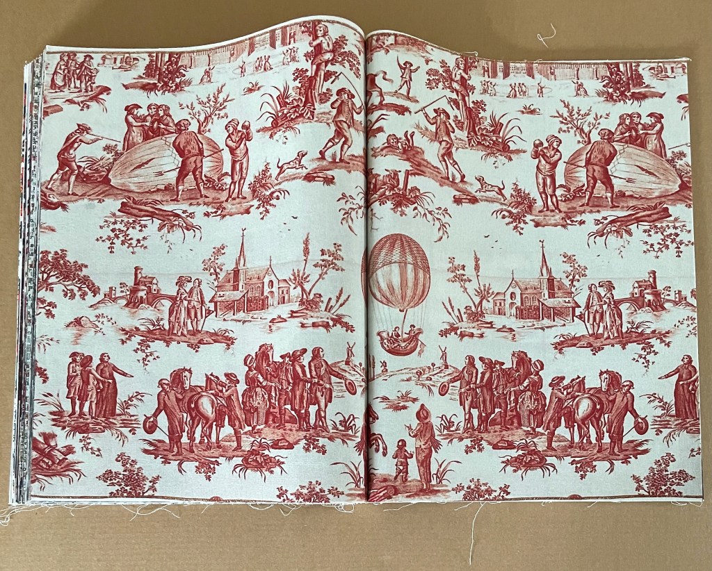 An open book featuring a vintage textile design with red and white illustrations depicting various scenes, including people, animals, and structures in a pastoral setting.