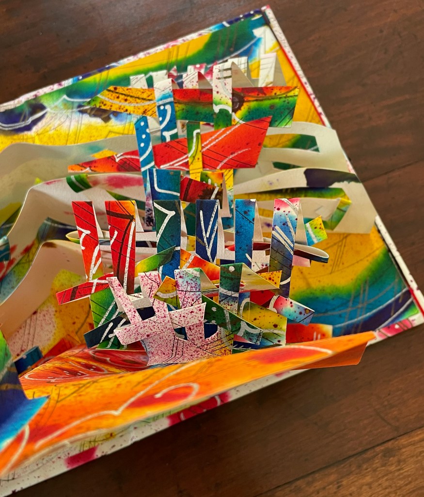A close-up view of a colorful pop-up art piece with various cut-out shapes in vibrant colors, set against a bright, abstract background.