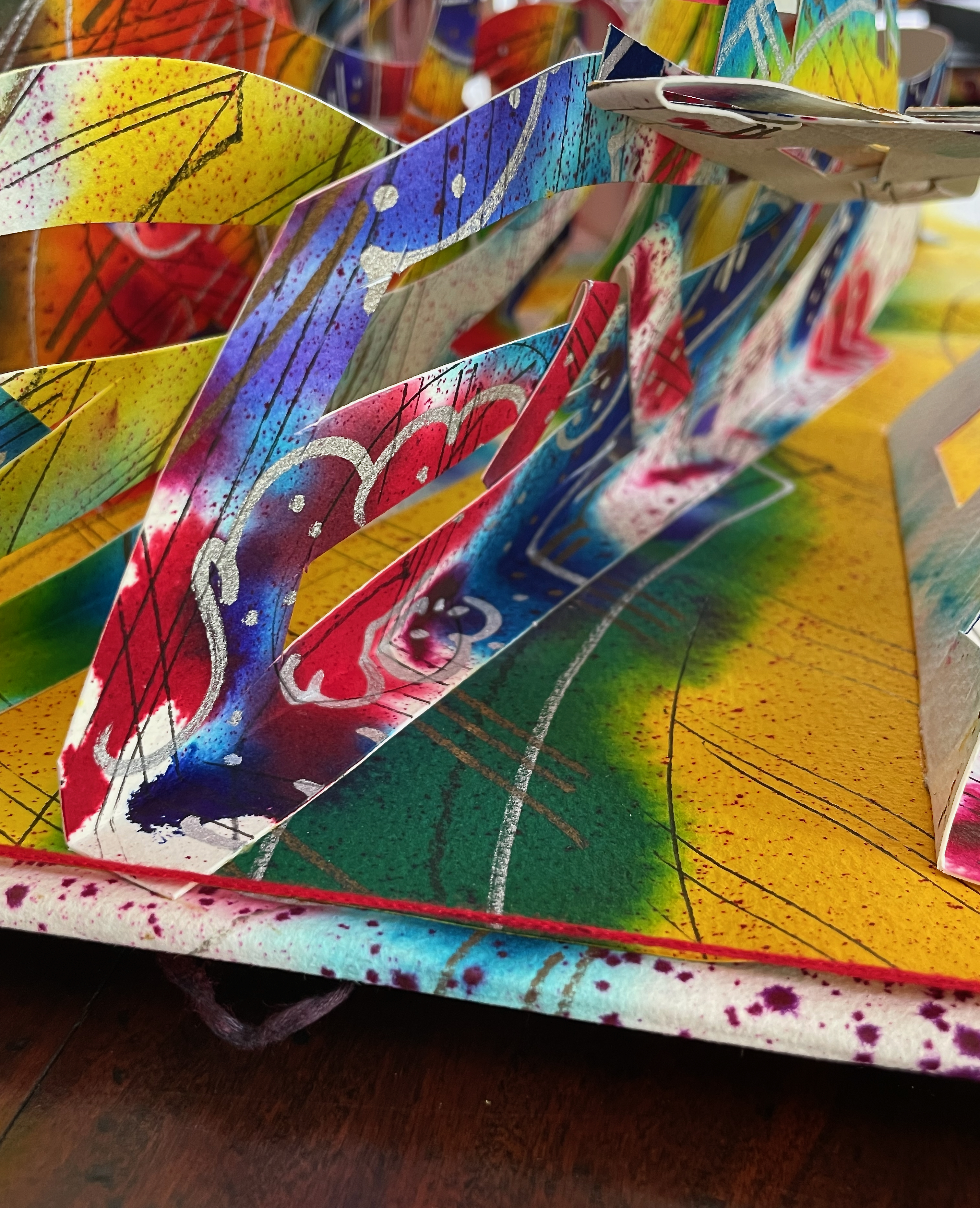 A close-up view of an intricate, colorful paper sculpture featuring vibrant patterns and textures. The artwork includes layered, cut-out designs with a mix of bright colors like red, yellow, blue, and green, creating a dynamic visual composition.
