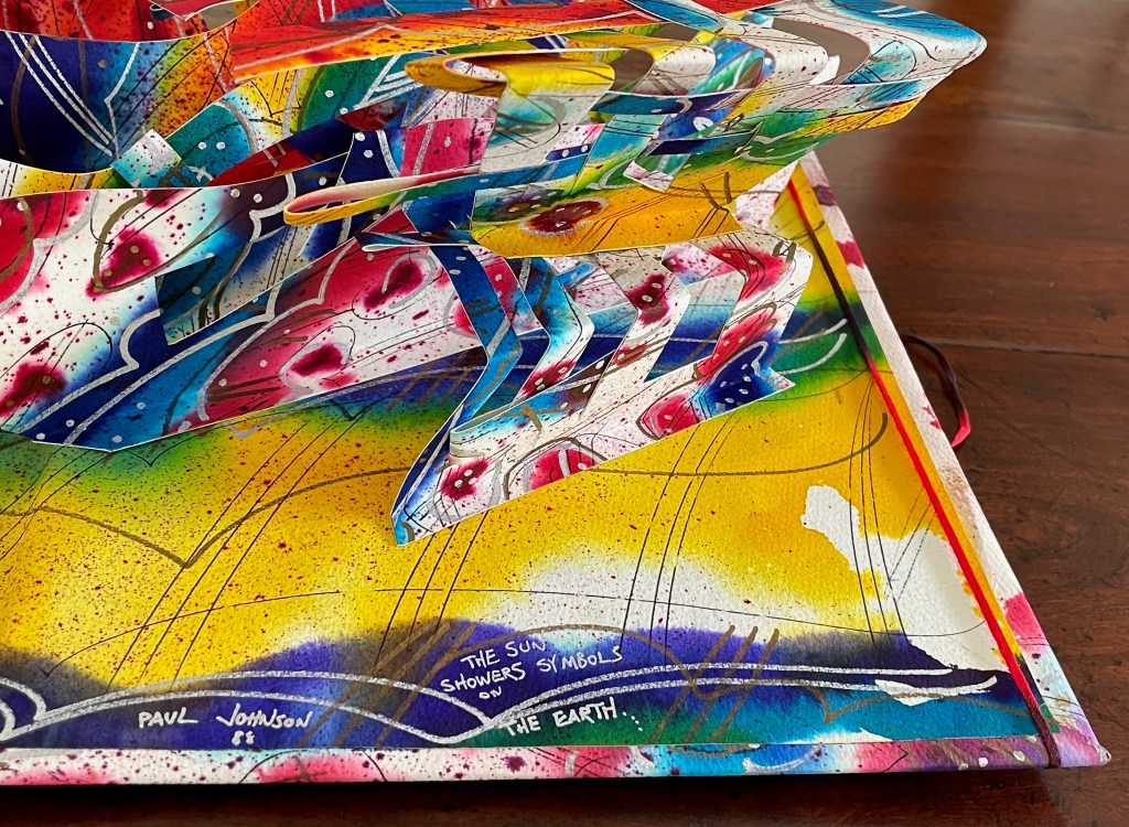 A colorful pop-up artwork featuring abstract shapes and vibrant patterns, set against a yellow and blue background, with text including the name 'Paul Johnson' and the phrase 'The sun showers symbols on the Earth.'
