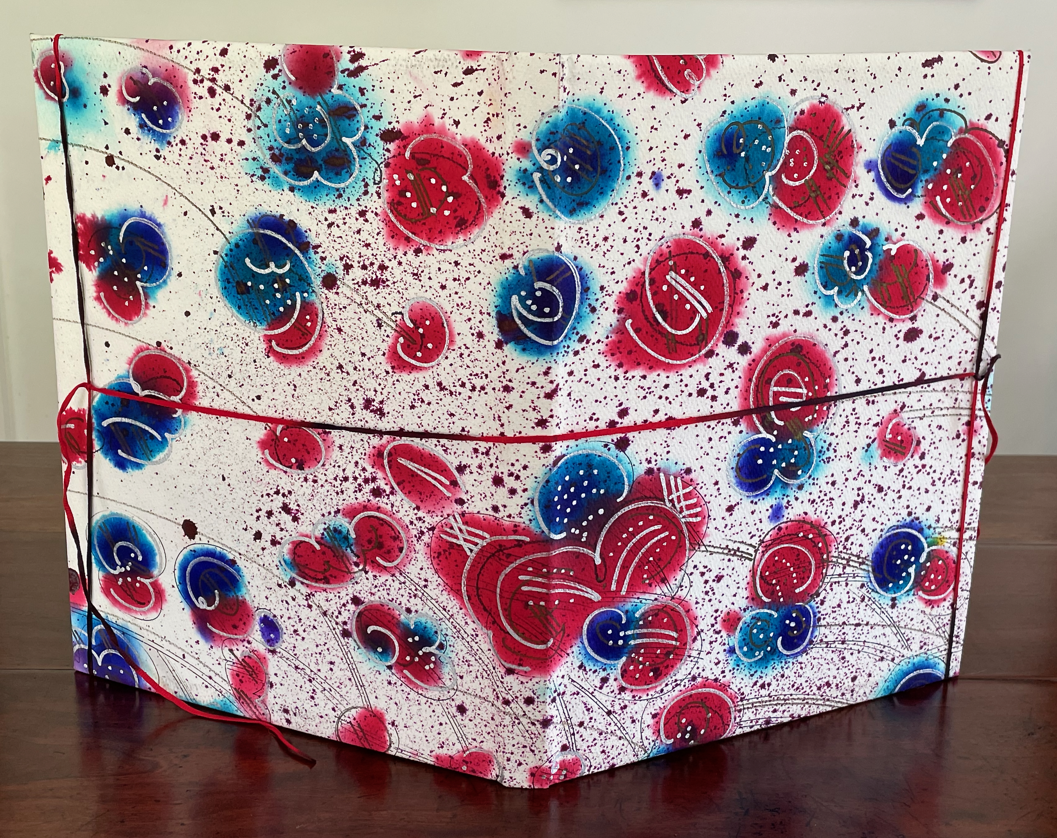 A decorative folder or binder featuring a colorful design with red, blue, and turquoise floral patterns on a white background, adorned with paint splatters.