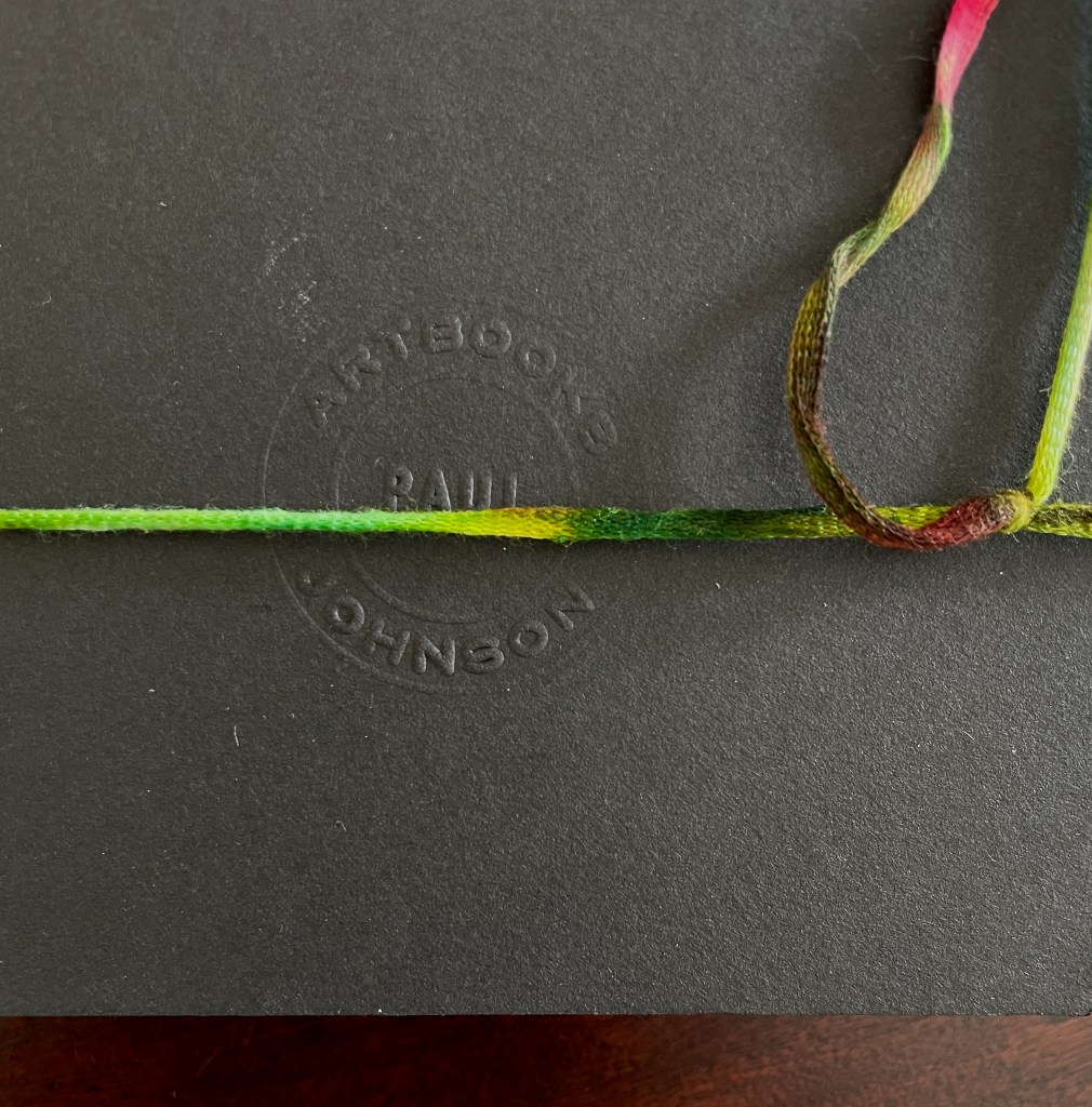 Close-up of a black book cover with an embossed text reading 'ARTBOOKS RAUL JOHNSON' and a colorful, multicolored string across it.