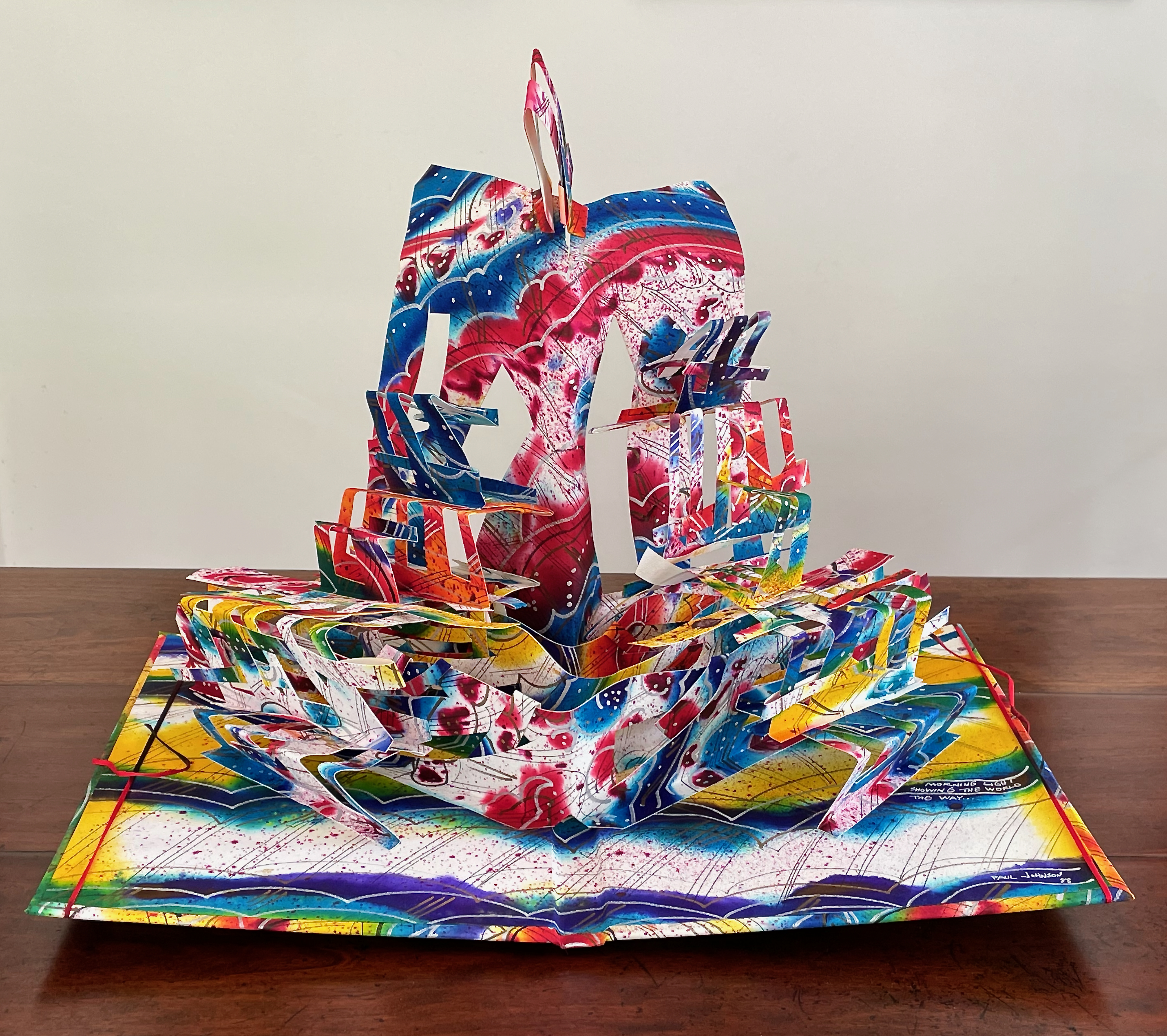 A colorful pop-up book page featuring a vibrant, intricately designed ship with layered sails, set against a bright abstract background.