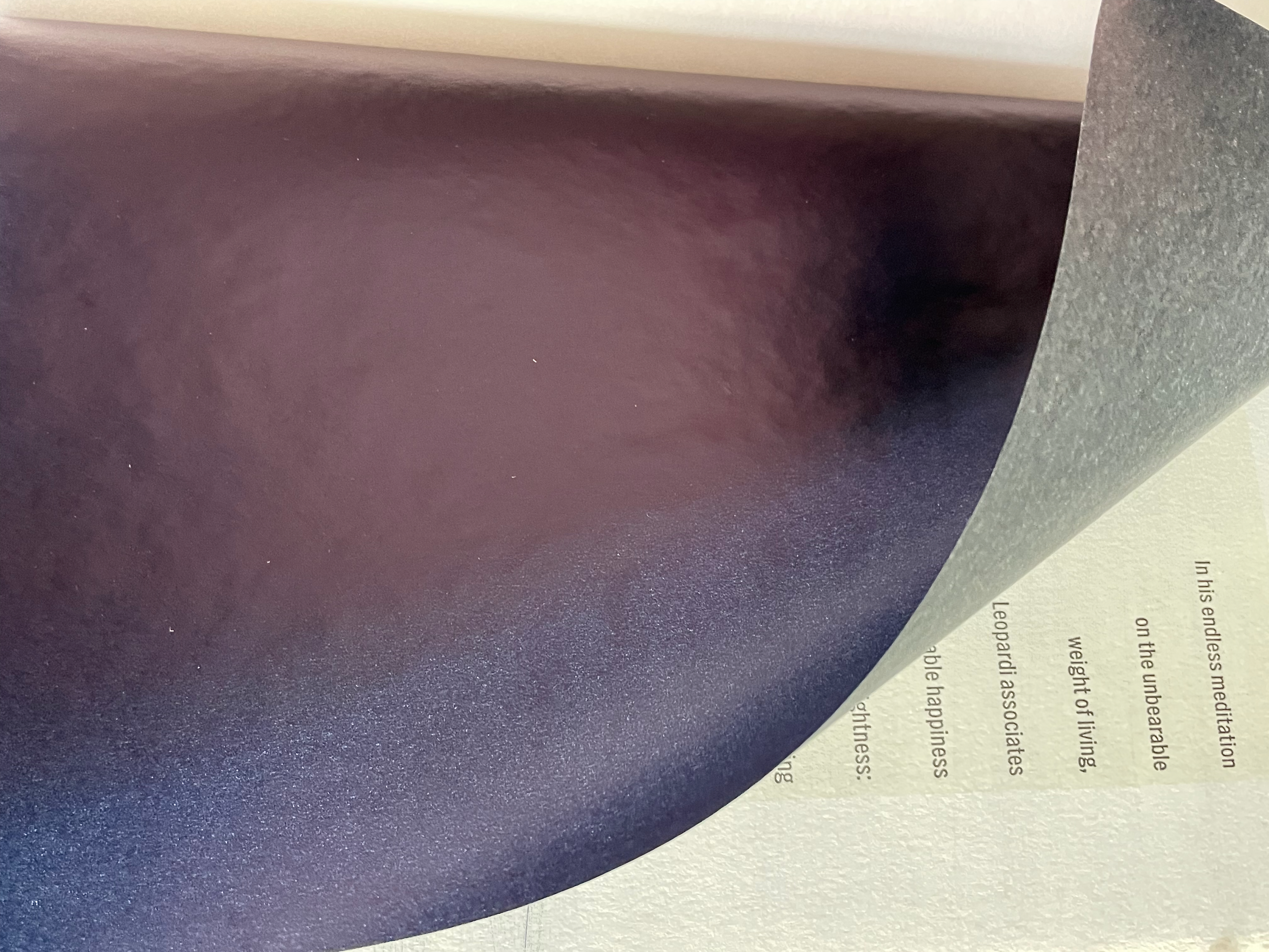 A close-up of a glossy, dark purple page being turned, revealing a sheet of white paper with text partially visible.