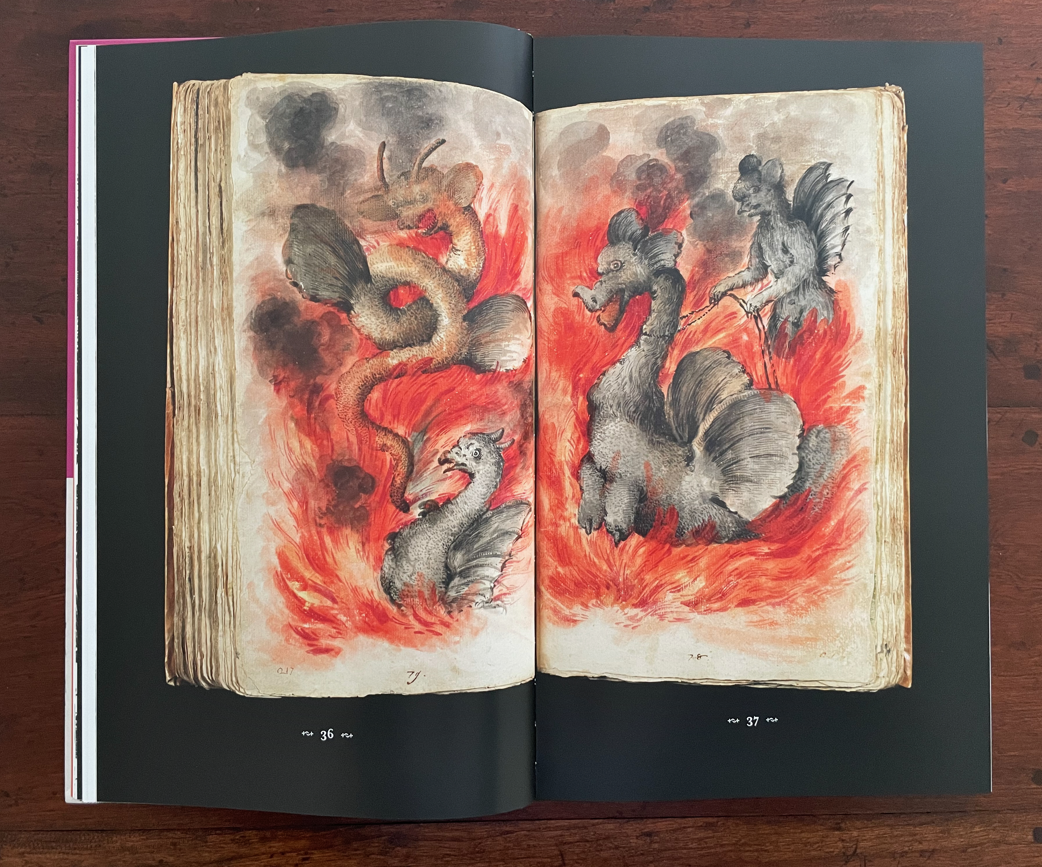 An open book displaying an illustration of fantastical creatures amid flames, with a dragon-like figure coiling, a bird-like creature, and a humanoid figure interacting with a large, mythical beast.