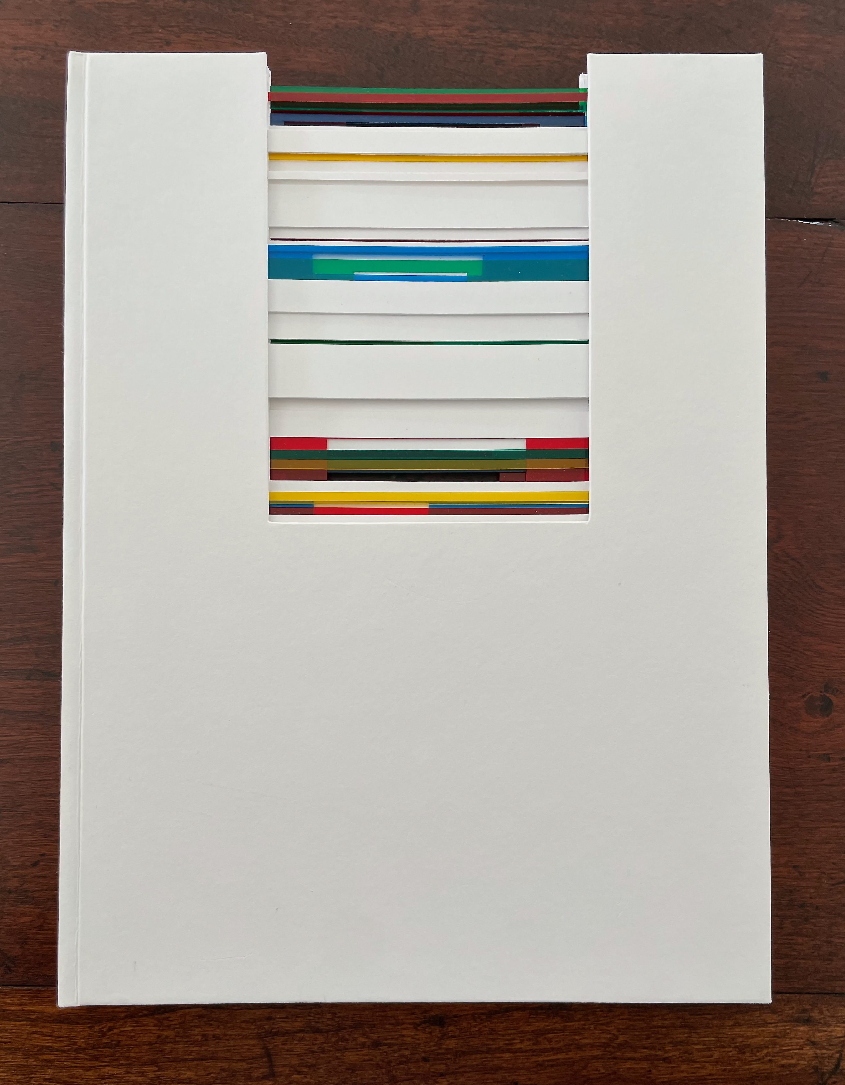 A white hardcover book with a cut-out on the front revealing colorful pages inside, arranged in a stacked pattern.
