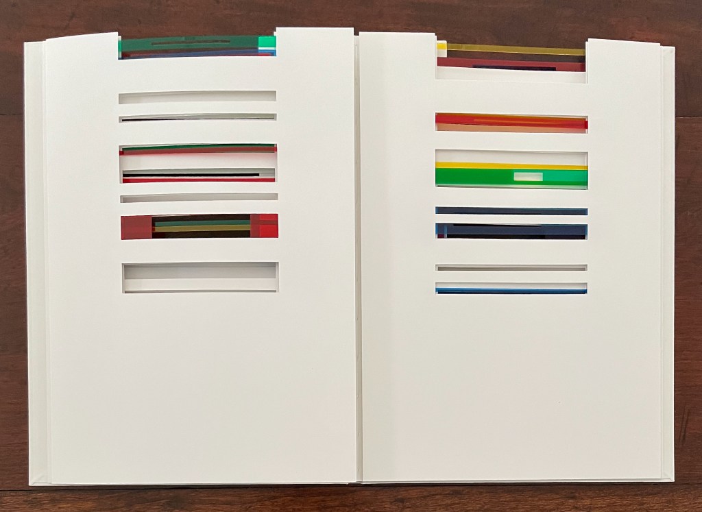 An open book with white pages featuring colorful strips inserted in various slots, showcasing a playful arrangement of translucent colors.