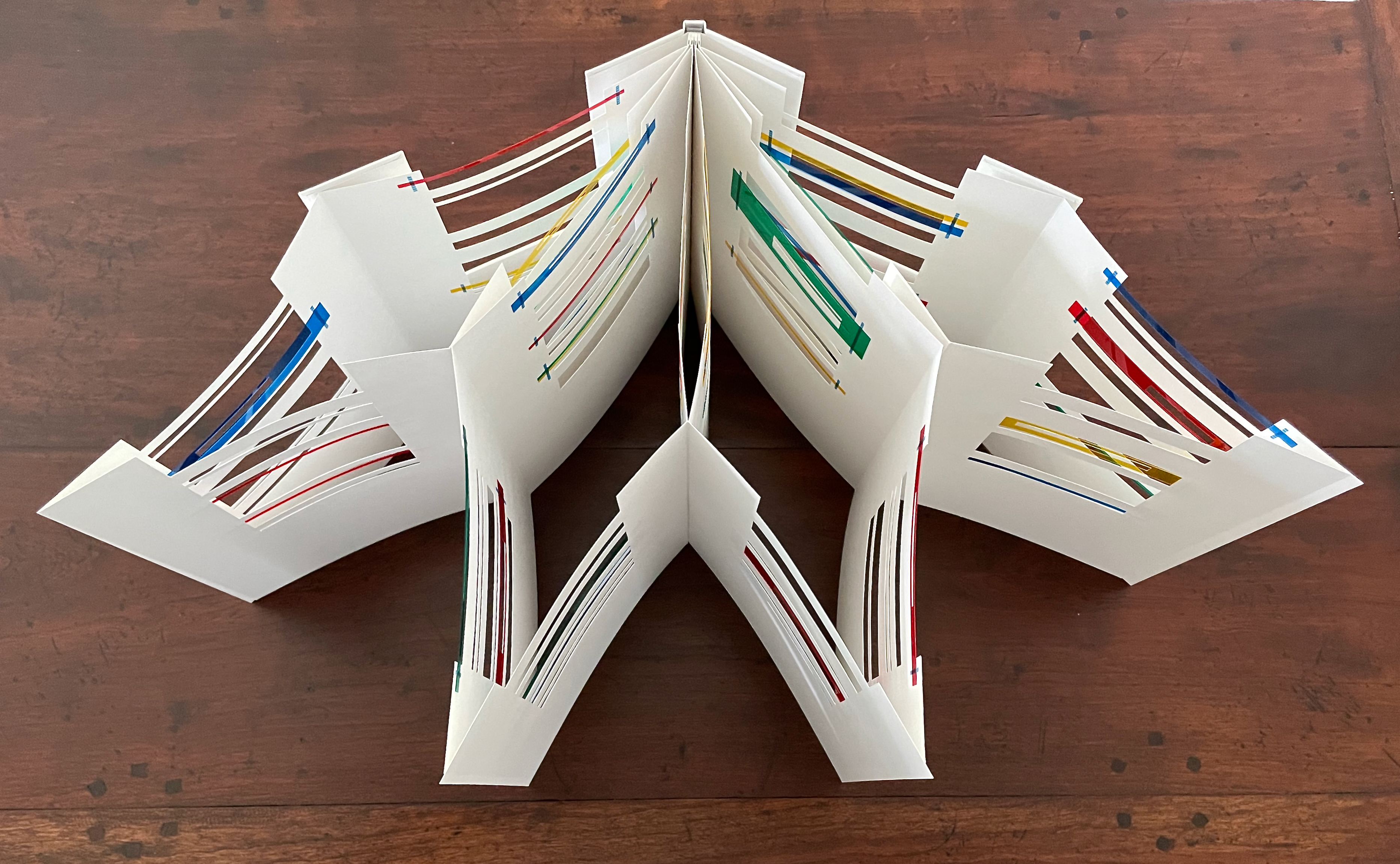 A handcrafted book structure made of white paper, featuring colorful strips of paper interwoven between the pages, displayed on a wooden surface.