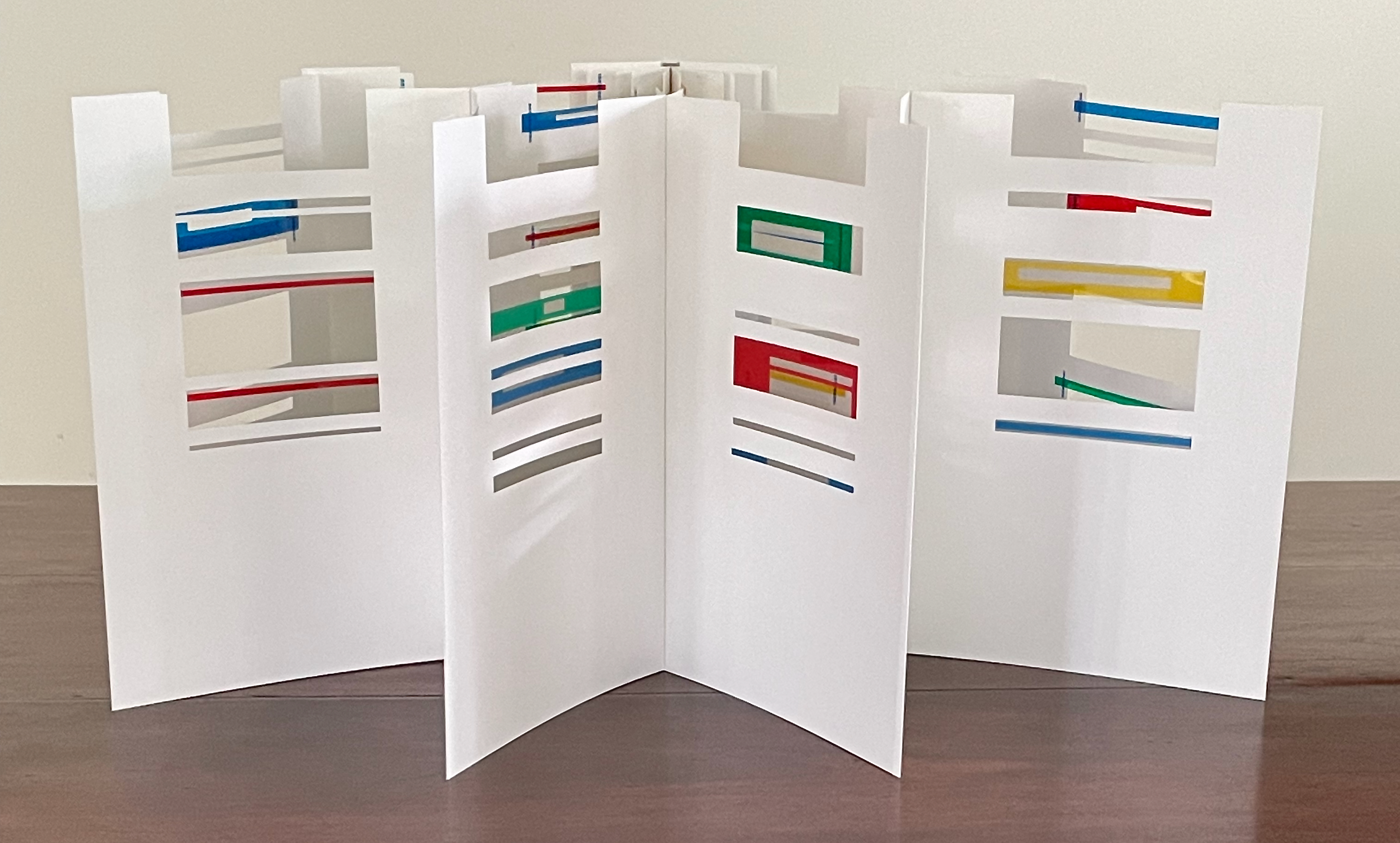 A white paper display featuring multiple sections with cut-out designs and colored strips in red, green, yellow, and blue, arranged on a wooden surface.