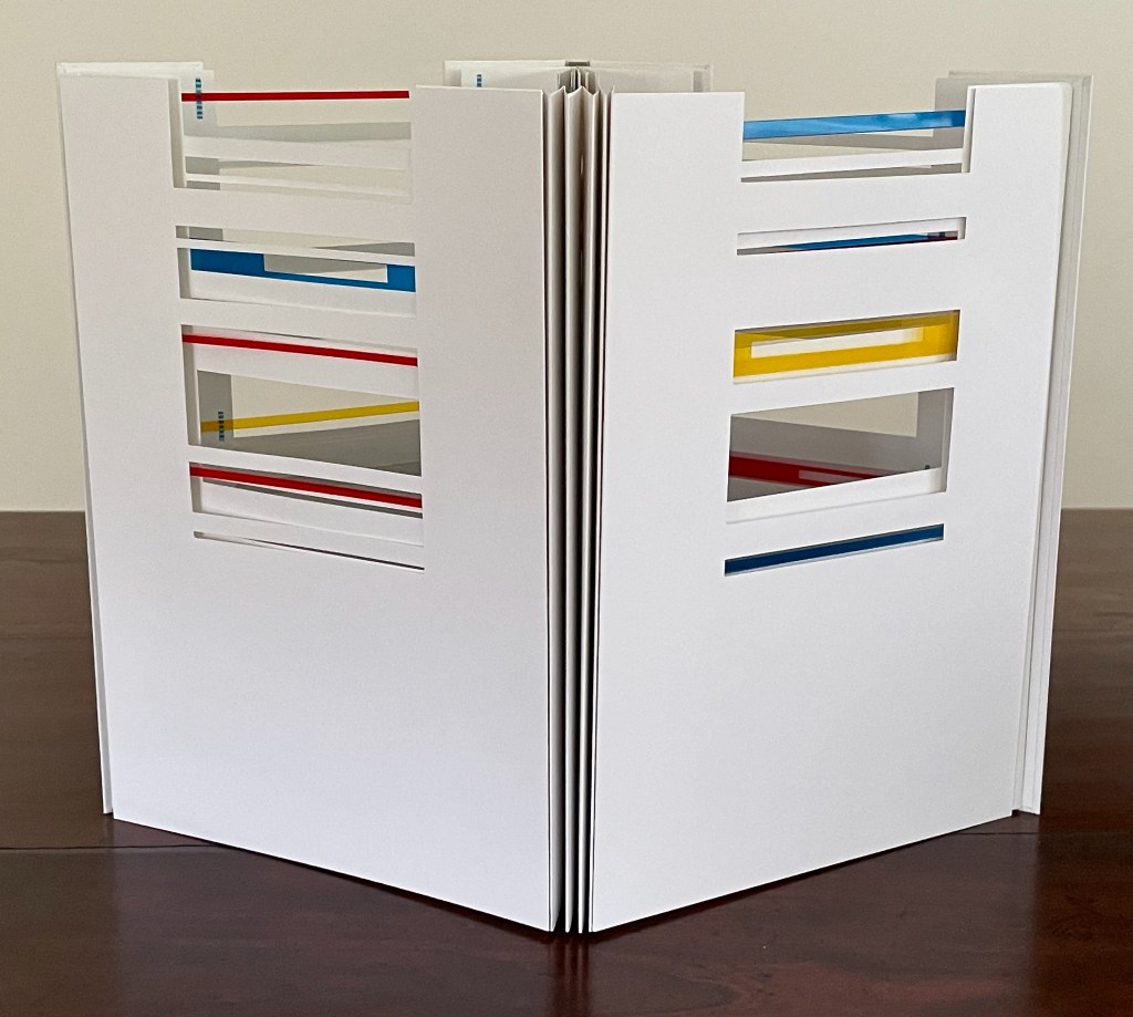 A close-up view of an open book with a white cover, featuring colorful cut-out patterns in red, blue, yellow, and white on its pages.