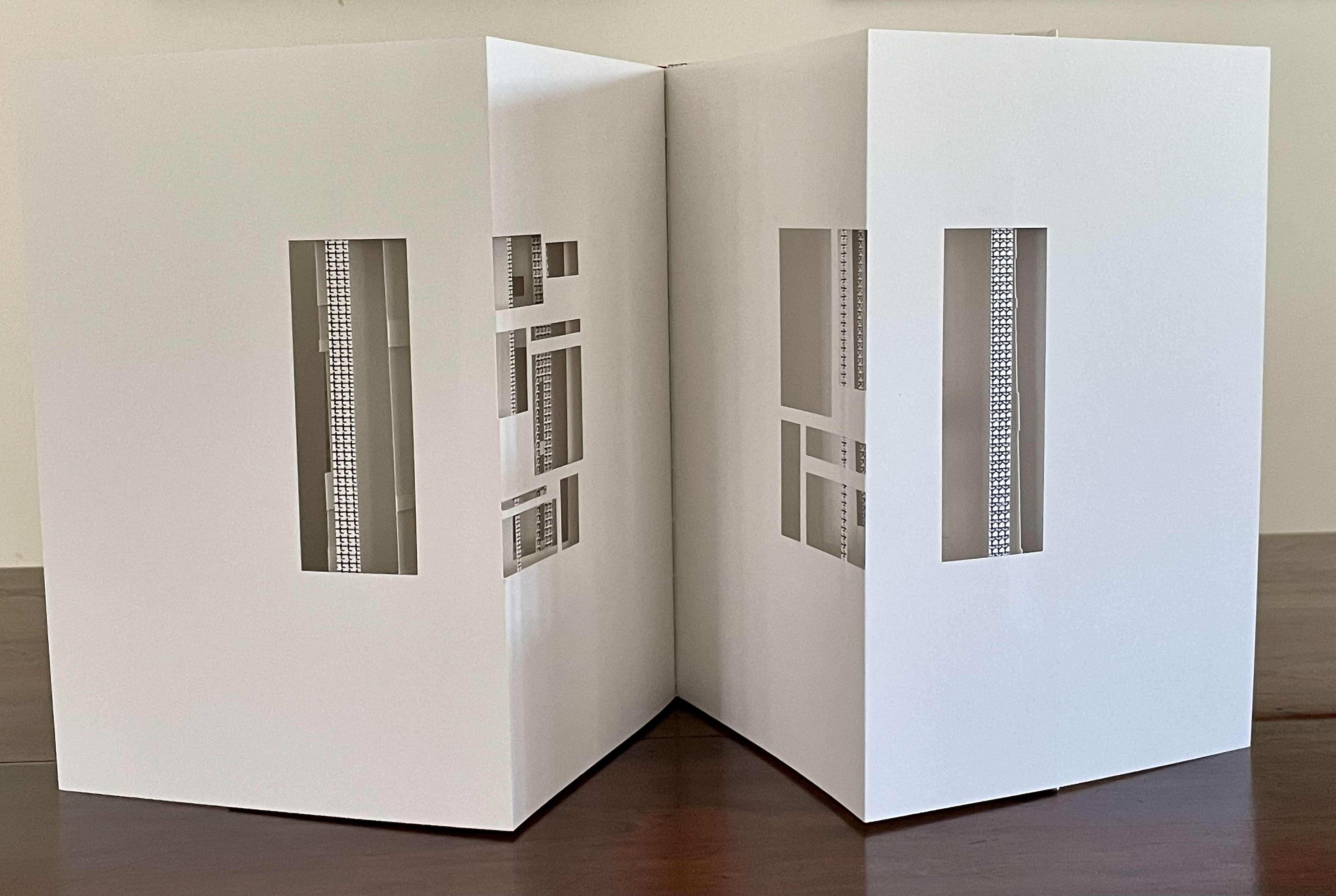A split architectural model made of white paper, featuring cut-out windows on either side, positioned on a brown wooden surface.