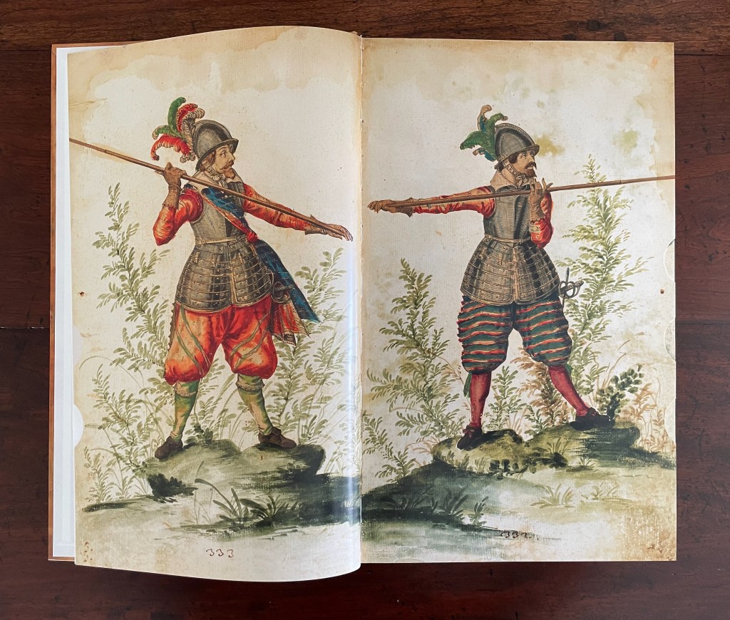 A historical illustration depicting two male soldiers wearing ornate armor and vibrant clothing, each holding a spear. They stand on rocky terrain with greenery in the background.