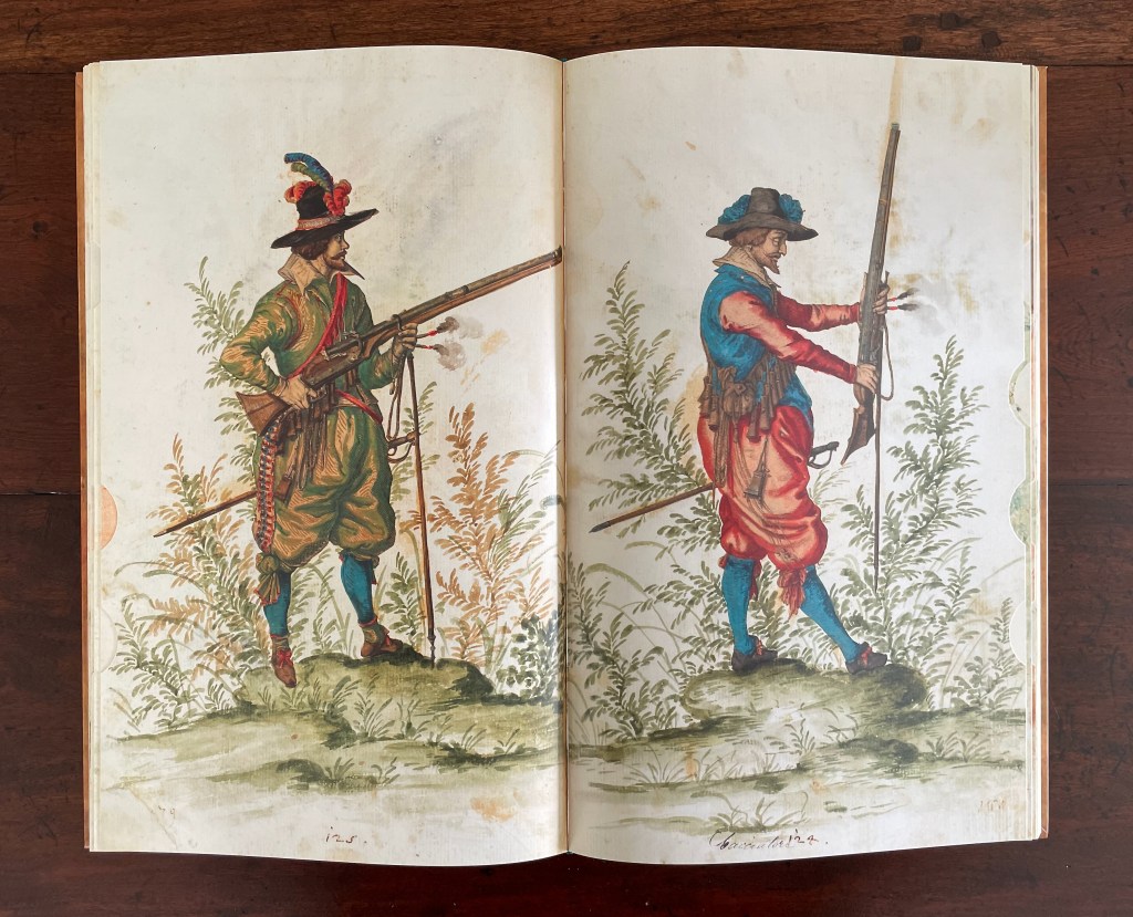 Two men in historical costumes, each holding a firearm, standing amidst greenery. The man on the left wears a green outfit with a feathered hat and a belt, while the man on the right is dressed in blue and red, with a more relaxed posture.
