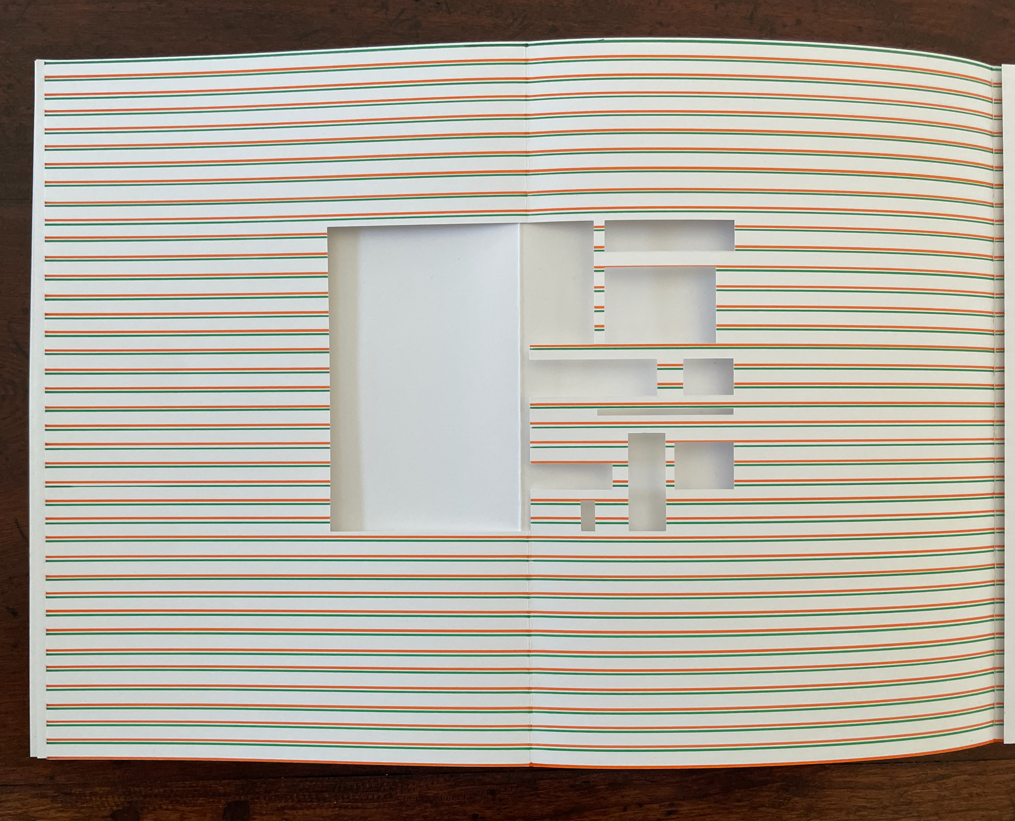An open book displaying a page with horizontal lines in white, green, and orange. The page features several cut-outs in various rectangular shapes, revealing a blank interior area.