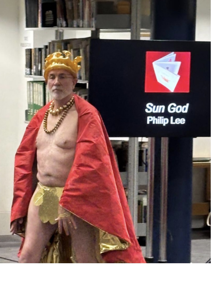 A man dressed as a king wearing a golden crown, a red cape, and a gold skirt, posing in front of a sign that reads 'Sun God' and 'Philip Lee'.