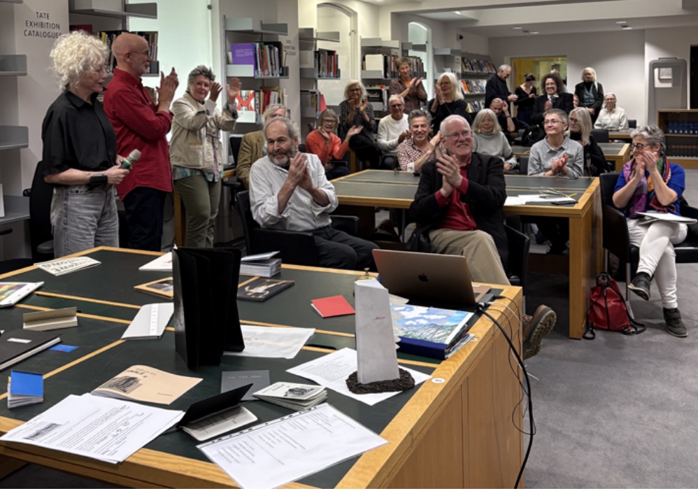 A group of people applauding in a library setting, with several tables displaying books and materials. The audience shows diverse reactions, and a few individuals are standing while others are seated.